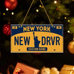 May include: A rectangular ornament resembling a New York license plate. The ornament is yellow and blue, with the words "NEW YORK", "NEW DRVR", and "CUSTOM NAME" printed on it. The Statue of Liberty is featured in the center. The ornament is hanging from a gold string.