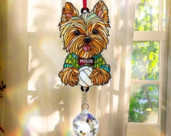 May include: A stained glass suncatcher depicting a Yorkshire Terrier dog wearing a green sweater with the name "Molly" on it. The suncatcher is suspended from a red ribbon and has a crystal prism hanging beneath it.