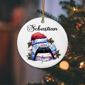 May include: A white ceramic Christmas ornament with the name "Sebastian" in elegant script. The ornament features a festive design of a video game controller wearing a Santa hat, surrounded by holly and Christmas lights. The year "20XX" is displayed on the controller.