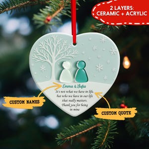 May include: A heart-shaped ornament with a light blue background, featuring a white tree and snowflakes. Two figures are depicted, one white and one teal. Text includes "Emma & Sofia" and a custom quote area. The ornament is made of ceramic and acrylic.
