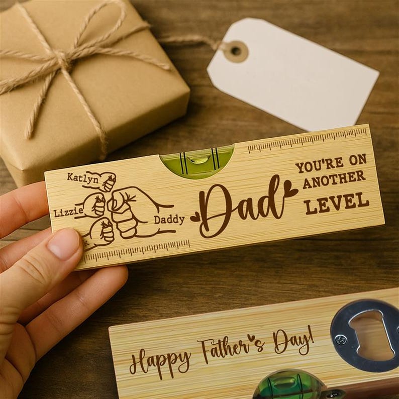 Pu&ograve; includere: Una livella in legno con la parola "Dad" e la frase "You're on Another Level" incise. Presenta una grafica di tre pugni e i nomi "Katlyn" e "Lizzie".