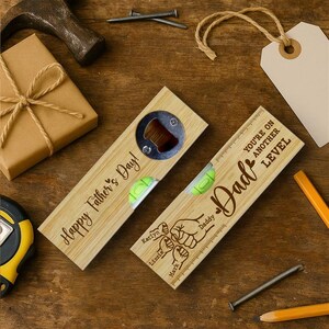Pu&ograve; includere: Due apribottiglie in legno con livelle a bolla e messaggi per la festa del pap&agrave;. Uno dice "Happy Father's Day!" e l'altro "Dad" con "You're on another level" e nomi. Sono visibili anche una confezione regalo, un martello, chiodi e matite.