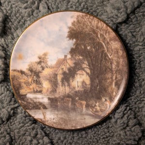 May include: A round decorative plate with a landscape scene. The plate features a detailed painting of a river, a house, trees, and figures in a boat. The plate has a gold-colored rim.