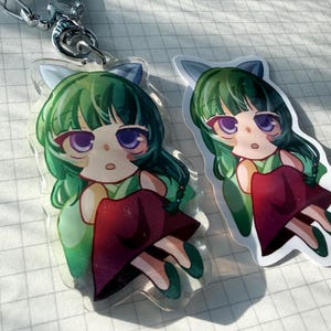 Op de afbeelding: Een doorzichtige acryl sleutelhanger en een bijpassende sticker met een chibi-stijl personage met lang groen haar, paarse ogen en katachtige oren. Het personage draagt een groen topje en een donkerrode rok en zit in een zittende houding.