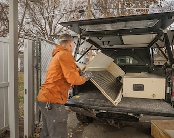 May include: A person is loading a wooden drawer and a wooden panel into the back of a truck. The truck has a black canopy with a window. The truck is parked in a driveway.