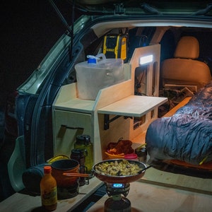Könnte beinhalten: Der Rücksitz eines Autos wurde in einen Campingplatz mit einer Holzplattform, einem Schlafsack und einer kleinen Küchenzeile mit einem tragbaren Herd und Kochutensilien umgewandelt.