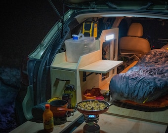 May include: A car's backseat is converted into a camping space with a wooden platform, a sleeping bag, and a small kitchen area with a portable stove and cooking utensils.