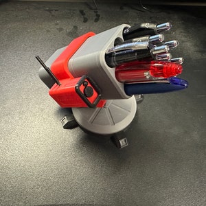 Könnte beinhalten: Ein grauer und roter 3D-gedruckter Stifthalter in Form eines Geschützturms mit drehbarer Basis. Der Halter hat mehrere Fächer für Stifte und Bleistifte. Der Halter ist mit verschiedenen Stiften gefüllt, darunter schwarze, blaue und rote Stifte.