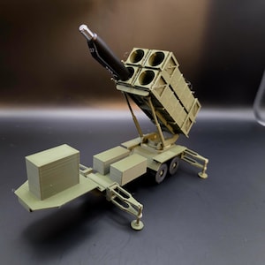 May include: A detailed model of a military missile launcher, painted in olive green. The launcher features a rectangular base with wheels, a raised missile compartment with four openings, and a black missile. The model is on a dark surface.