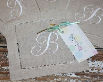 May include: Set of four linen napkins with embroidered letter B in a script font. The napkins are beige and the embroidery is white. The napkins are stacked on top of each other and a tag with the text "Rozados" is attached to the top napkin.