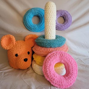 May include: A handmade crochet stacking toy with a central white post and colorful rings in blue, pink, orange, yellow, and teal. An orange bear head is next to the toy, and two additional rings in blue and purple are also present.