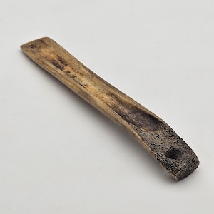 May include: A handcrafted, elongated wooden incense holder. The holder is light brown with a dark brown, textured end and a small hole. The trough is carved out, and the overall design is simple and natural.