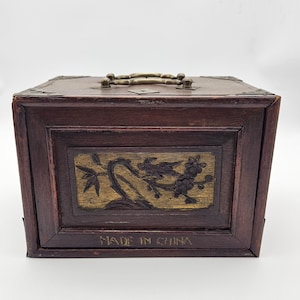 May include: A rectangular dark brown wooden box with a brass handle on top and brass corner accents. The front panel features a recessed gold-toned rectangular carving of a bird on a branch. The text MADE IN CHINA is printed on the front.