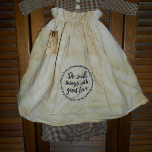 May include: A handmade, rustic-style apron and top set. The top is a plaid pattern in brown, blue, and red. The apron is off-white with a circular text design that reads "Do small things with great love". A small tag is attached.