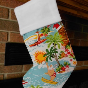 May include: Christmas stocking with a white cuff and a tropical print. The print features Santa surfing, palm trees, a tiki, and a reindeer surfing. The background is a light blue with a bright yellow sun and red flowers.