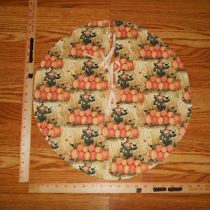 May include: A round tree skirt with a fall harvest theme. The skirt features a repeating pattern of pumpkins, scarecrows, and hay bales in shades of orange, green, and beige. The skirt is tied with a light pink ribbon.