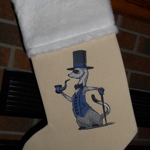 May include: Beige Christmas stocking with a white cuff. The stocking features an embroidered meerkat wearing a blue vest, top hat, and holding a pipe and cane. A festive holiday decoration.