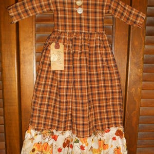 May include: A hanging fabric dress ornament featuring a brown and black plaid pattern with two white buttons at the neckline. A rectangular tan tag with the handwritten text Harvest Time is attached, and the hem has a white ruffle with pumpkins.