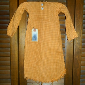 May include: Orange and white checkered fabric dress with long sleeves, two blue buttons, and a frayed hem. A small tag with a black and white image hangs from the dress. The dress is hanging against a wooden shutter background.