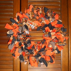 May include: A handmade wreath with a festive autumn theme. The wreath is constructed from strips of fabric in orange, black, and plaid patterns. The wreath is circular and appears to be hanging on a wooden door.