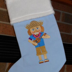 May include: Light blue Christmas stocking with a white cuff. The stocking features an embroidered image of a nutcracker playing a ukulele, wearing a straw hat, blue shirt, and khaki shorts. A festive holiday decoration.