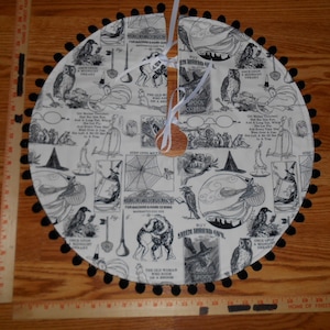 May include: A round, cream-colored Halloween tree skirt with a black pom-pom trim. The skirt features a repeating black and white vintage illustration pattern with witches, owls, and text. A wooden tree trunk opening is at the center.