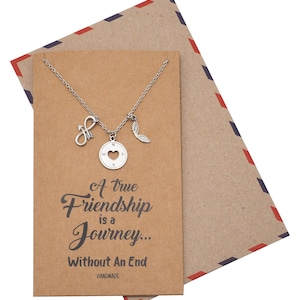 May include: A silver necklace with three charms: an arrow, a compass with a heart cutout, and a wing. The necklace is displayed on a brown card with the text "A true Friendship is a Journey... Without An End". An envelope is behind the card.