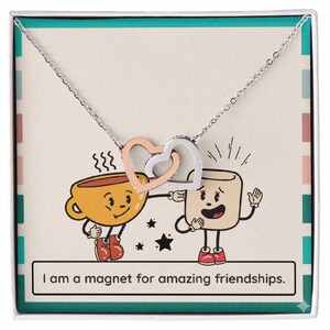 Könnte beinhalten: Eine silberne Halskette mit zwei ineinandergreifenden Herzanhängern, einer roségoldenen und einer silbernen, präsentiert auf einer Karte mit einer Cartoon-Kaffeetasse und einem Marshmallow. Auf der Karte steht: "Ich bin ein Magnet für tolle Freundschaften."