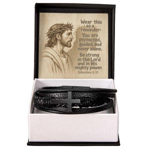 May include: Black leather bracelet with a black cross charm, displayed in a white box with a black frame. The box lid features a drawing of Jesus with text that reads "Wear this as a reminder: You are protected, guided, and never alone."