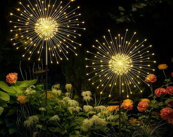 May include: Two solar-powered garden lights shaped like bursts of light. The lights are composed of many small, warm white LED lights radiating outwards. The lights are set in a garden with flowers and greenery, creating a decorative outdoor lighting display.