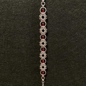 May include: A silver-colored bracelet with alternating sections of chainmail-style links and round, dark red beads. The bracelet has a lobster clasp closure and is displayed against a dark background.