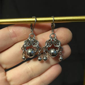May include: Silver-toned earrings with a chainmail design. Each earring features a large, round, polished metal bead at the center, surrounded by intricate chainmail. Three smaller beads dangle below. The earrings are displayed against a dark background.