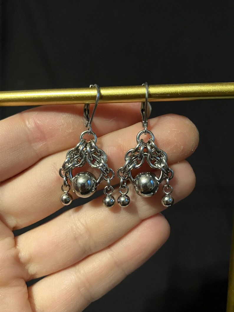 May include: Silver-toned earrings with a chainmail design. Each earring features a large, round, polished metal bead at the center, surrounded by intricate chainmail. Three smaller beads dangle below. The earrings are displayed against a dark background.