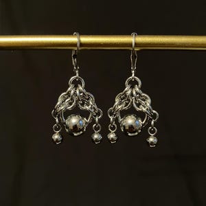 May include: A pair of silver-toned earrings. Each earring features a central sphere, surrounded by intricate chainmail-style metalwork. Three smaller spheres dangle below the central sphere, creating a layered effect. The earrings hang from a simple hook.