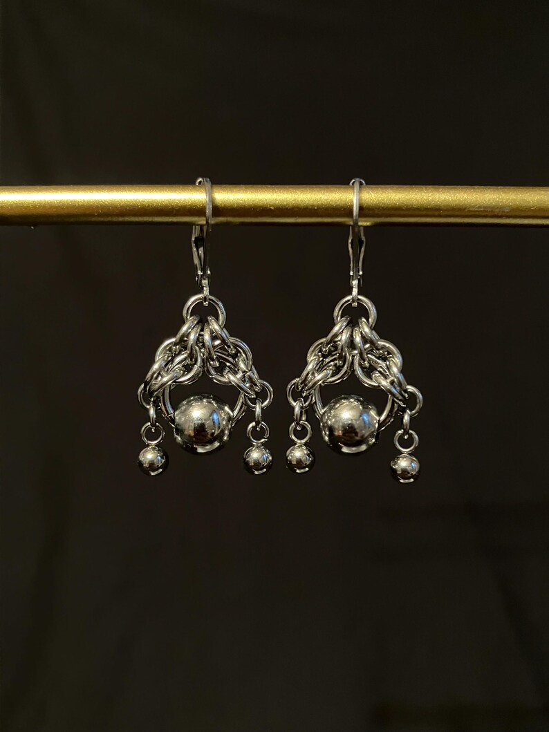 May include: A pair of silver-toned earrings. Each earring features a central sphere, surrounded by intricate chainmail-style metalwork. Three smaller spheres dangle below the central sphere, creating a layered effect. The earrings hang from a simple hook.