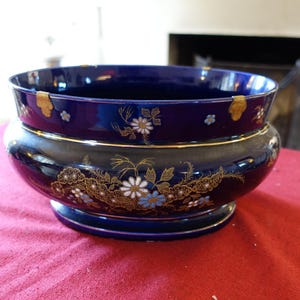 Peut inclure: Un grand bol en céramique bleu foncé avec des bordures dorées et des motifs floraux. Le bol a une forme large et arrondie avec une base en piédestal. Des fleurs blanches et bleues ornent l'extérieur.
