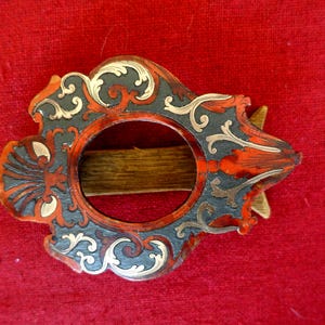 Peut inclure: Boucle décorative ornée avec une ouverture circulaire centrale. La boucle présente une base gris foncé avec des accents rouges et dorés, et des volutes complexes. Le fond est rouge uni.