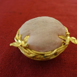 May include: A small, round, pink pincushion with a gold metal frame. The frame is decorated with a design of birds and leaves.
