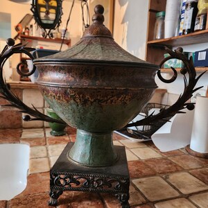 May include: A decorative metal urn with a lid, featuring a weathered green and bronze patina finish. The vessel has two ornate, leaf-shaped handles on either side and rests on a square, dark metal base with intricate scrollwork detailing.