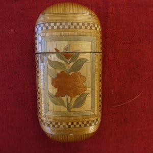 May include: A small, cylindrical wooden box with a floral design. The box is made of thin strips of wood that are woven together to create a pattern. The floral design is in shades of brown, orange, and green. The box is closed and has a hinged lid.