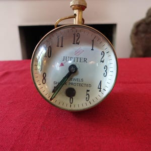Peut inclure: Une horloge sphérique vintage avec une tige en laiton et un cadran en verre transparent. Le cadran est blanc avec des chiffres noirs et le mot "JUPITER" imprimé dessus. L'horloge repose sur une surface rouge.