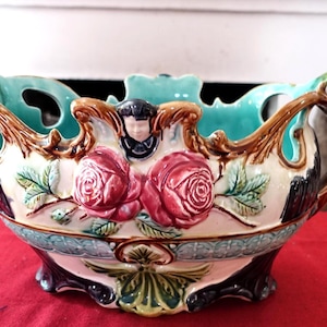 Puede incluir: Un cuenco decorativo de cerámica con interior y detalles exteriores turquesa. El cuenco presenta rosas en relieve, hojas y una pequeña figura. Las asas y el borde son marrones, y la base es una mezcla de colores.