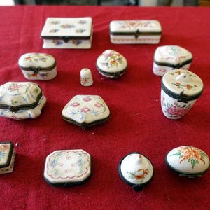 May include: A collection of 11 small, decorative porcelain boxes with floral designs. The boxes are in various shapes, including square, round, and oval. The boxes are all white with gold trim and feature intricate floral patterns in shades of pink, blue, and green.