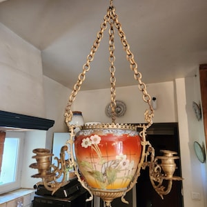 May include: A hanging chandelier featuring a central ceramic bowl with a gradient orange and yellow finish, decorated with painted white flowers and green leaves. The bowl is supported by ornate gold-toned metal chains and two candle holders.