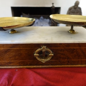 May include: A vintage balance scale featuring two brass pans mounted on a rectangular white marble top. The base is constructed from dark polished wood with a central circular brass emblem containing a small glass window and engraved text.