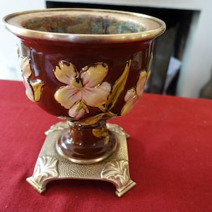 Peut inclure: Un vase en céramique marron avec un bord et une base dorés, orné de motifs floraux en relief crème et or. Le vase est posé sur un socle carré en métal orné, créant une présentation élégante.