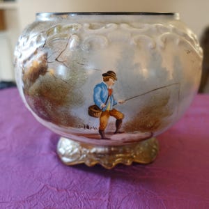 May include: A decorative glass bowl with a painted scene of a man fishing. The bowl has a rounded shape with a gold-colored base. The painted scene features a man in a blue jacket and brown pants, holding a fishing rod.