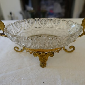 May include: A clear glass bowl with a decorative cut pattern featuring a scalloped rim. The bowl is supported by an ornate gold-toned metal base with three feet and two matching side handles, each displaying intricate scrollwork and detailing.