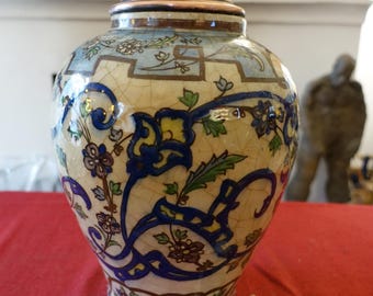 May include: A ceramic lamp with a copper top and a cream-colored base. The lamp is decorated with blue floral patterns and green leaves. The lamp has a white on/off switch and a yellow plug. The lamp is on a red surface.