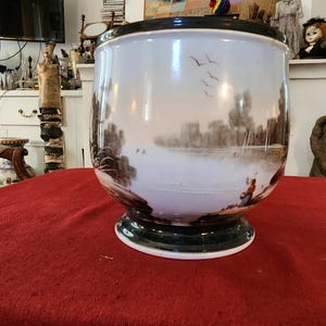 May include: A ceramic planter with a glossy white finish and a dark green rim and base. The exterior features a hand-painted landscape scene in sepia tones, depicting a calm lake, trees, flying birds, and a figure sitting near the water's edge.